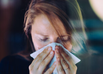 Woman sneezing behind a window, using a tissue.