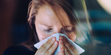 Woman sneezing behind a window, using a tissue.