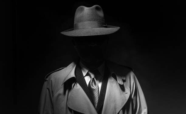 Man posing in the dark with a fedora hat and a trench coat, 1950s noir film style character